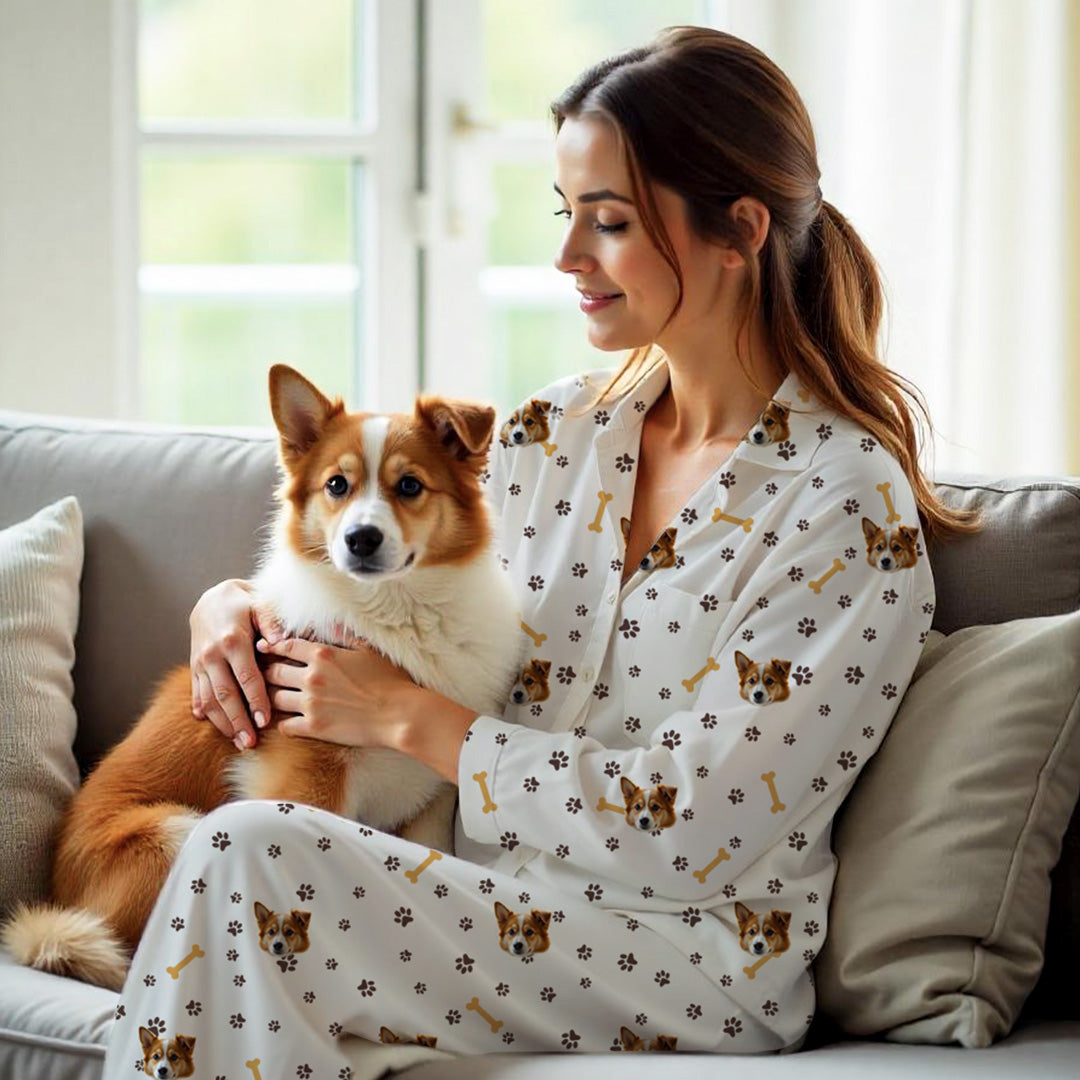 Woman wearing white custom pet pajama set with dog face, paw print, and bone design, holding brown and white dog on the sofa